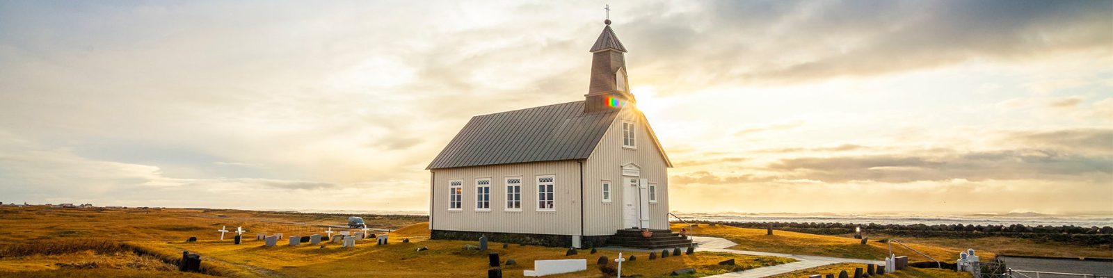 A small white church with a steeple stands alone in a grassy field under a dramatic sky at sunrise, gravestones scattered around and the sun peeking behind—an inspiring scene for religious organization printers capturing moments of faith.