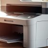 A modern, gray multi-function printer (MFP) sits on a desk in a well-lit office with shelves and a window with blinds in the background.