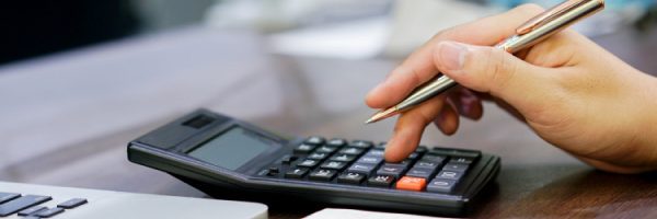 A person holding a pen uses a calculator next to an open laptop on a wooden desk, reviewing tariffs and business printing needs.