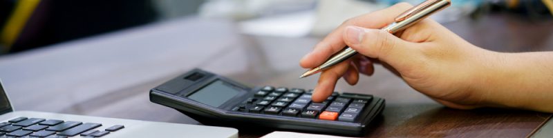 A person holding a pen uses a calculator next to an open laptop on a wooden desk, reviewing tariffs and business printing needs.