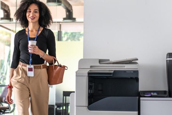 A woman with curly hair, holding a cup and a brown bag, smiles as she walks through an office hallway past some of the best copiers and a printer.