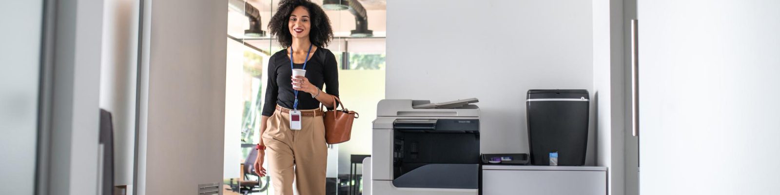 A woman with curly hair, holding a cup and a brown bag, smiles as she walks through an office hallway past some of the best copiers and a printer.