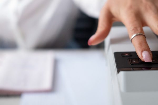 A person with manicured nails and a ring presses a button on a photocopier, often used by construction businesses, with paperwork and a pen visible on the desk in the background—highlighting the importance of commercial printers.
