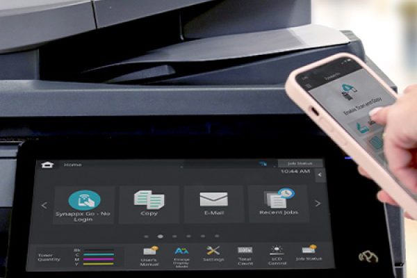 A person in a blue shirt uses a smartphone near a modern office copier with a touchscreen display showing printing and scanning options, ideal for businesses seeking professional printers.