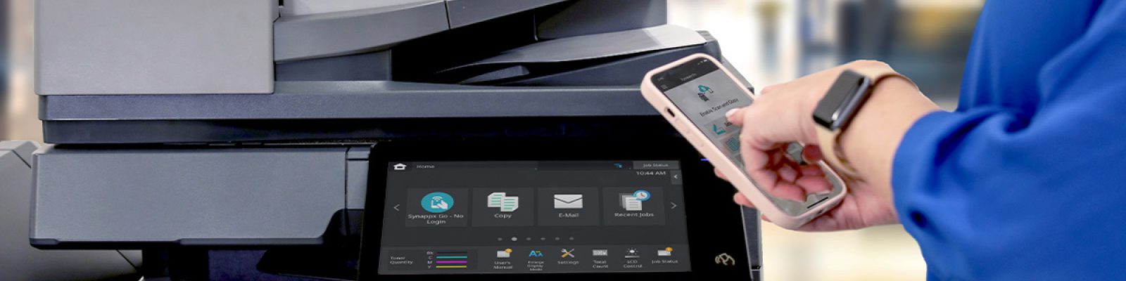 A person in a blue shirt uses a smartphone near a modern office copier with a touchscreen display showing printing and scanning options, ideal for businesses seeking professional printers.