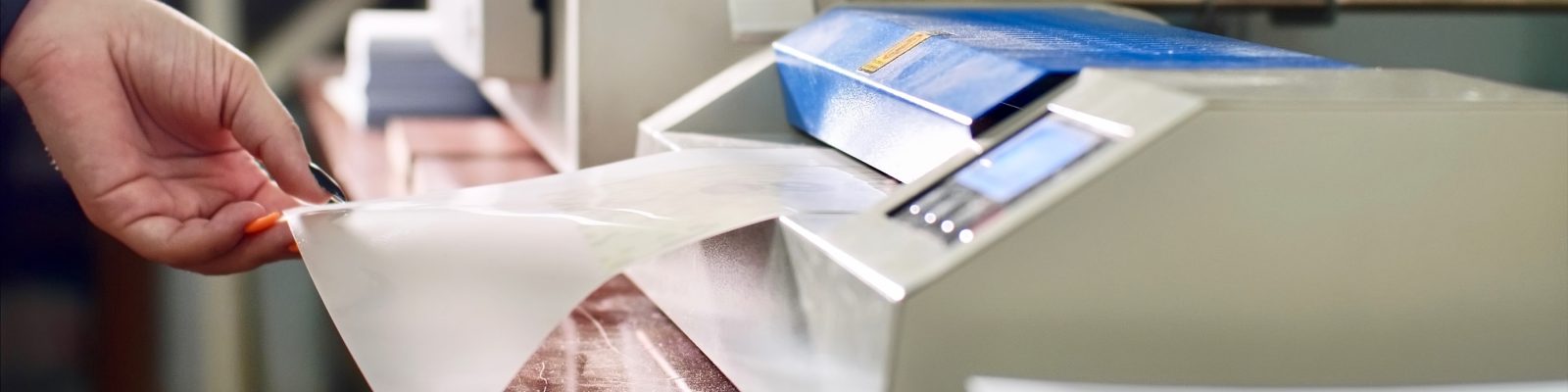 A close-up of a person’s hand feeding a sheet of paper into a laminating machine, as the machine covers the paper with clear plastic film for added document protection.