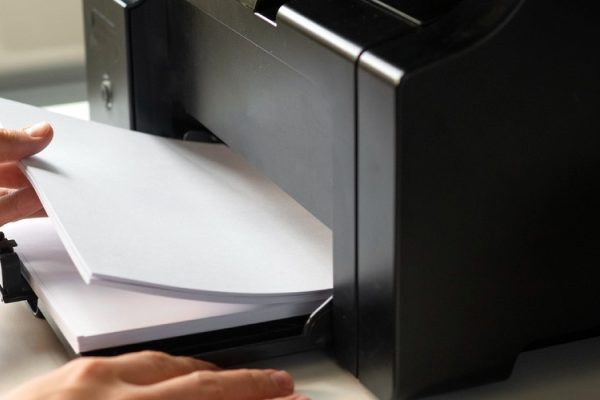 A person loads paper into a black printer—part of a managed print fleet—on a desk next to a computer monitor displaying folders.