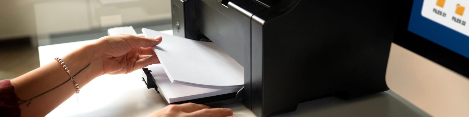 A person loads paper into a black printer—part of a managed print fleet—on a desk next to a computer monitor displaying folders.