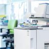 A modern office printer and copier sits in a bright, open-plan workspace—perfect for busy medical offices—with office chairs, desks, file folders, and partitions visible in the background.