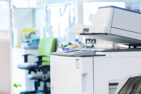 A modern office printer and copier sits in a bright, open-plan workspace—perfect for busy medical offices—with office chairs, desks, file folders, and partitions visible in the background.