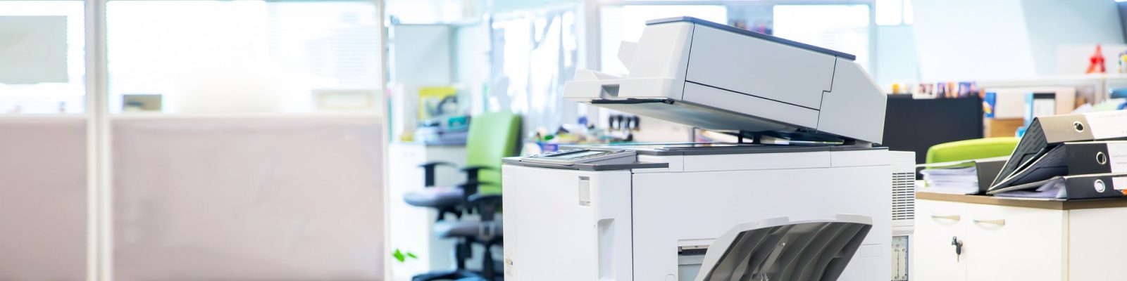 A modern office printer and copier sits in a bright, open-plan workspace—perfect for busy medical offices—with office chairs, desks, file folders, and partitions visible in the background.