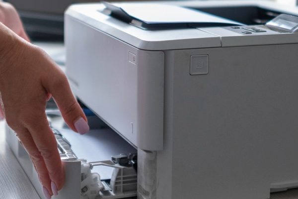A person in a blue jacket is opening the paper tray of a white all-in-one printer on a desk, possibly to add or remove paper. A keyboard and another printer are visible in the background.
