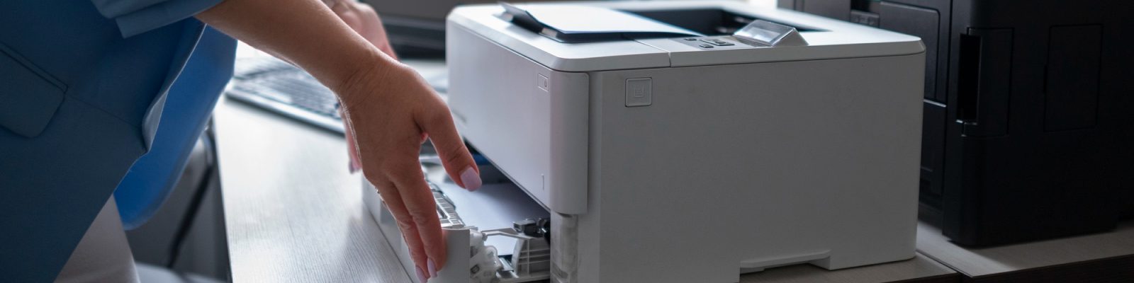 A person in a blue jacket is opening the paper tray of a white all-in-one printer on a desk, possibly to add or remove paper. A keyboard and another printer are visible in the background.