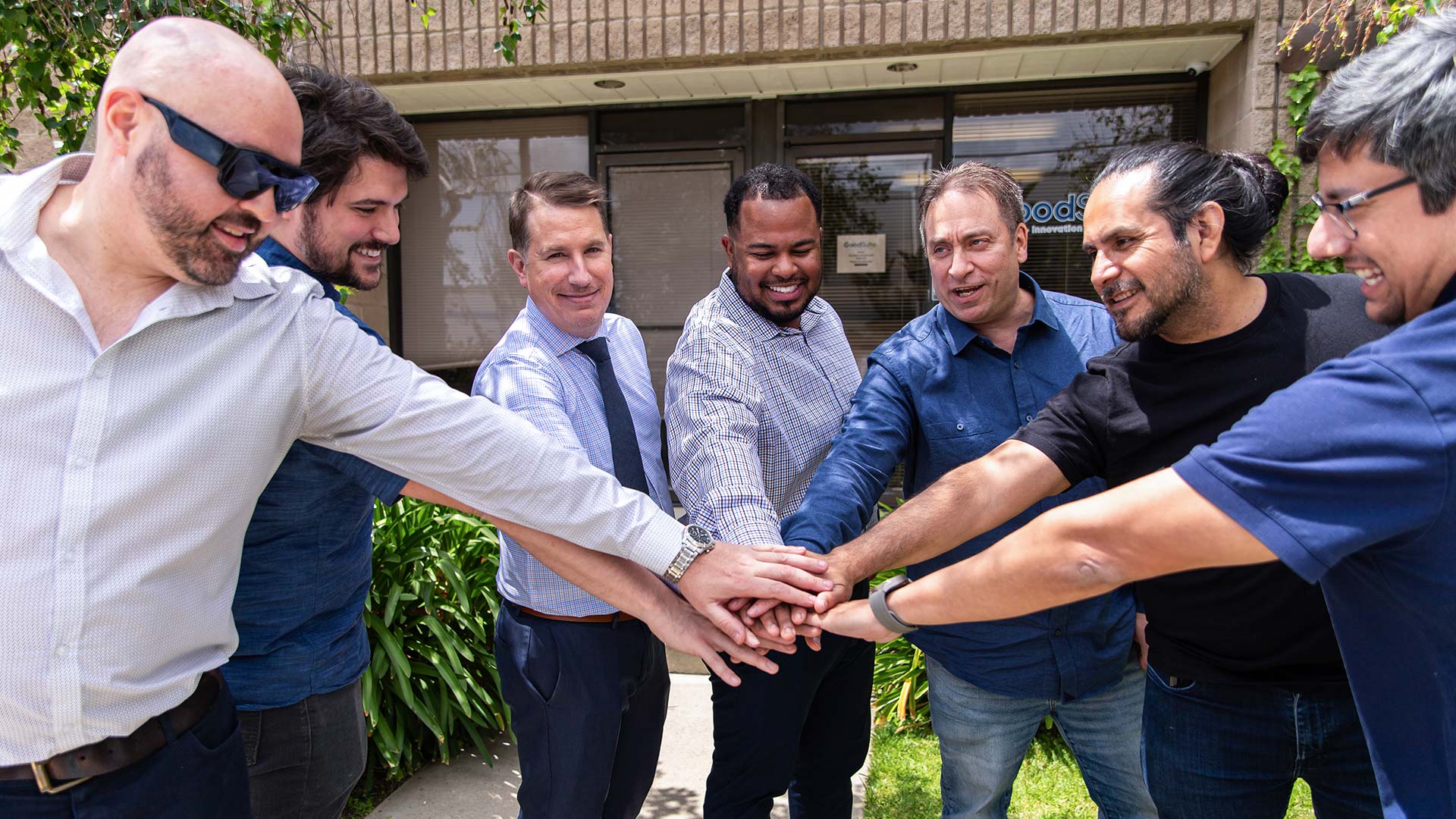 Seven men stand outdoors in a semi-circle, reaching their hands into the center for a group huddle, smiling and looking at each other in a show of teamwork and unity—reflecting the collaborative spirit at GoodSuite.