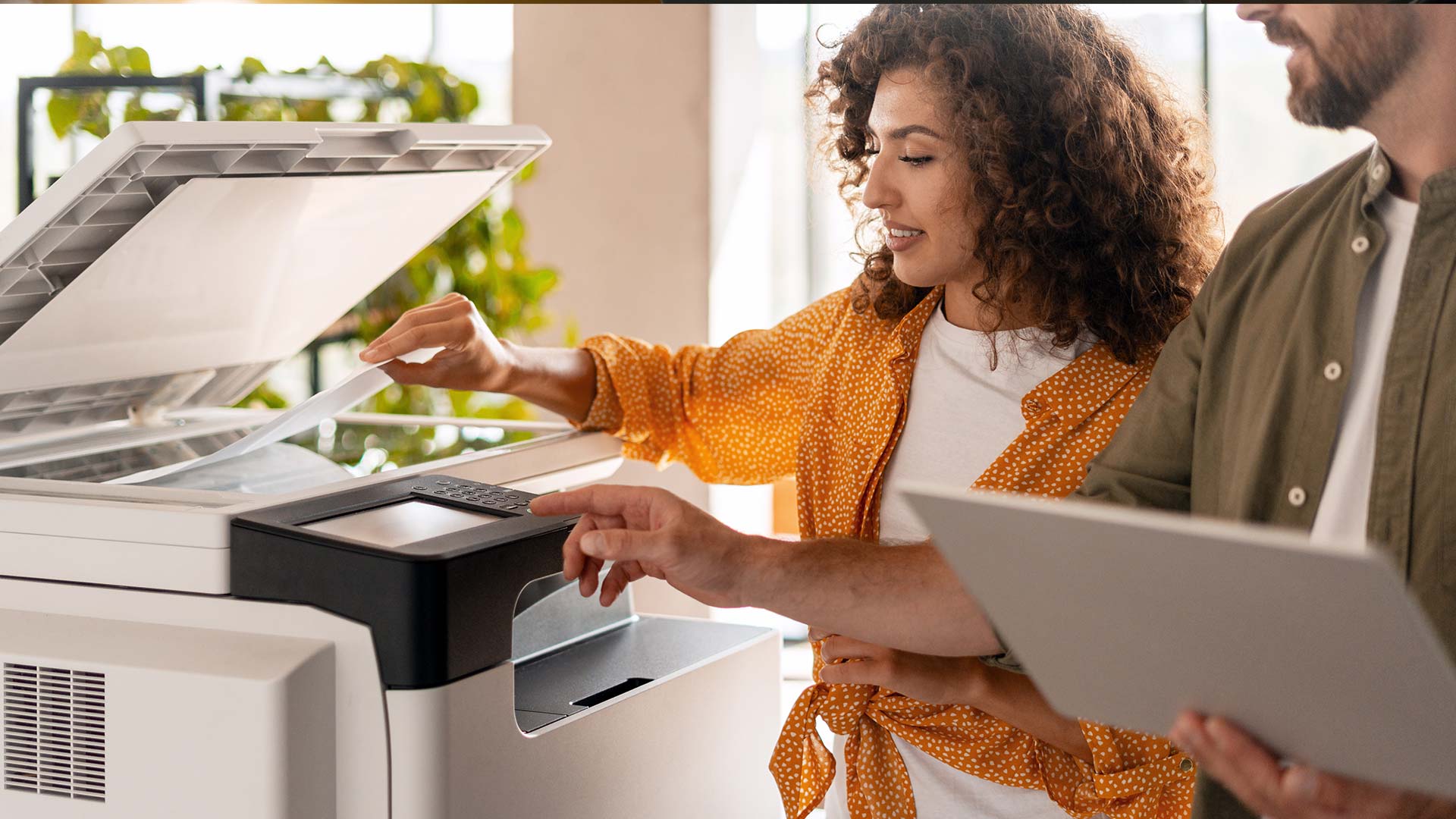 Two colleagues use a copier; a woman lifts the lid while a man holds a tablet and operates the machine’s touchscreen. Bright office setting with plants in the background, highlighting modern copiers in action.