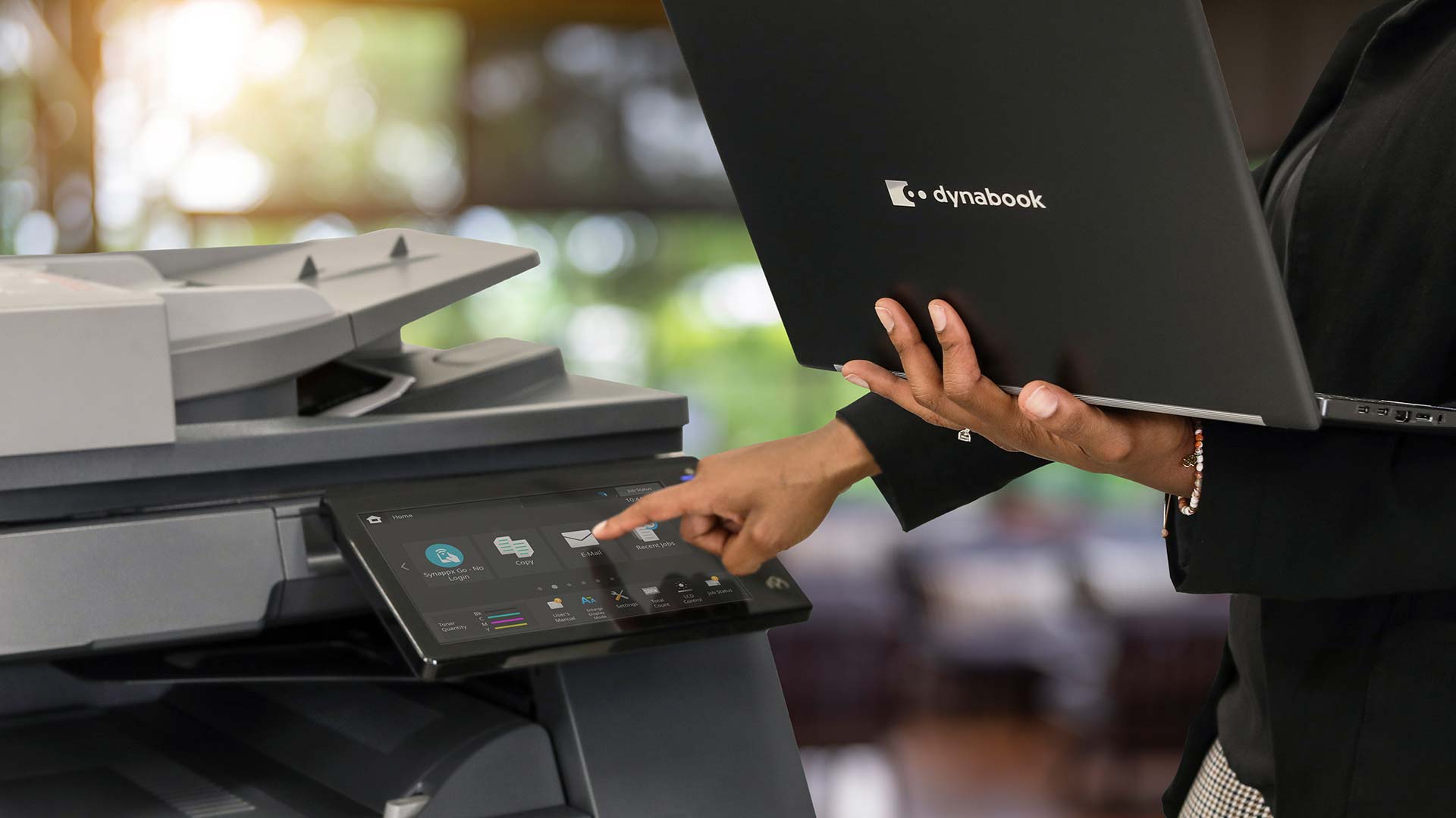 A person uses one hand to touch the touchscreen of a copier while holding an open Dynabook laptop in the other hand. The office setting, with blurred background, highlights multitasking with copiers and printers.