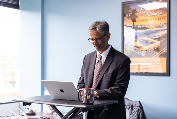 A man in a suit and tie works at a standing desk with a laptop and coffee mug in a modern office. A colorful poster with a car and palm trees hangs on the light blue wall, embodying the GoodSuite difference maker spirit.
