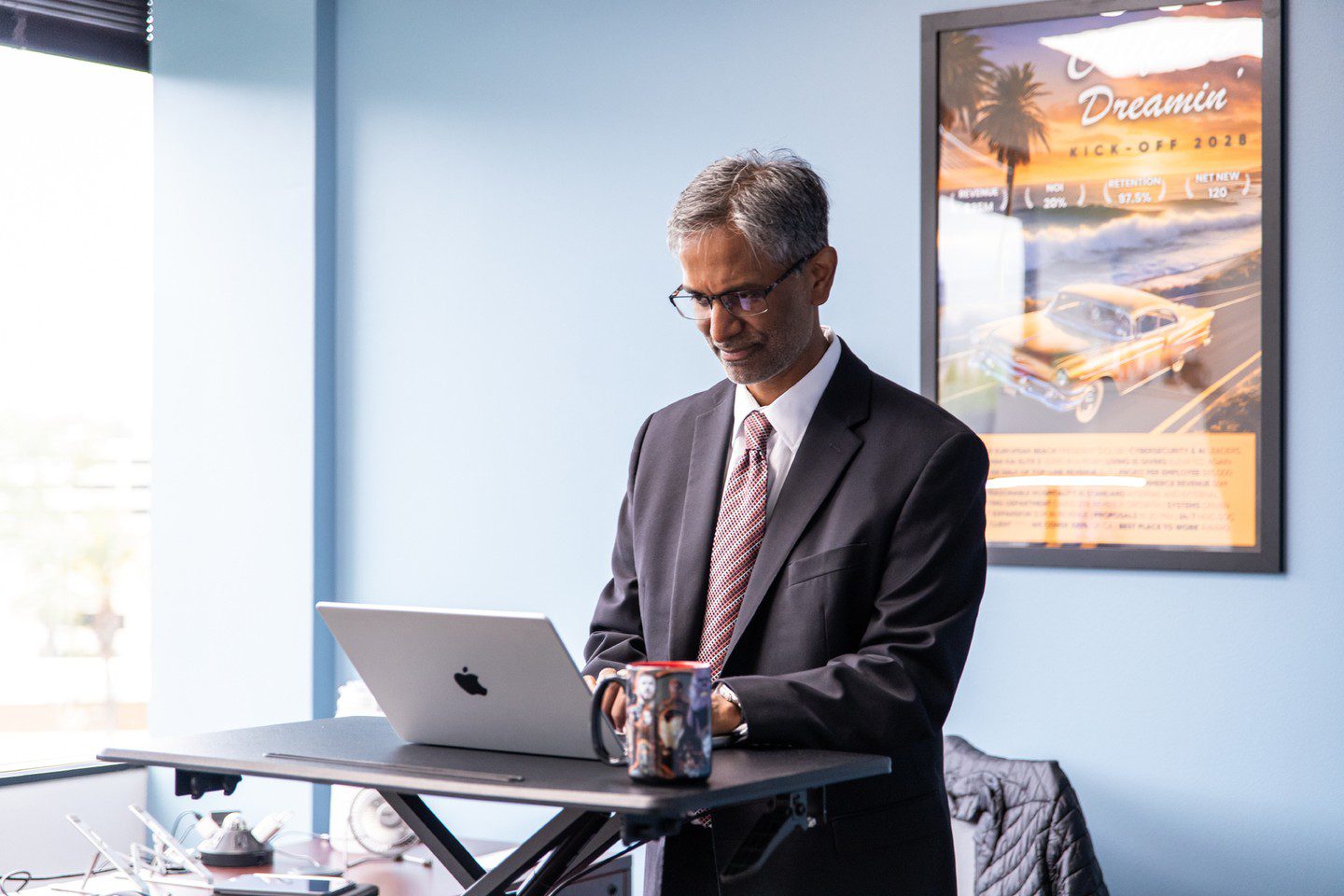 A man in a suit and tie works at a standing desk with a laptop and coffee mug in a modern office. A colorful poster with a car and palm trees hangs on the light blue wall, embodying the GoodSuite difference maker spirit.