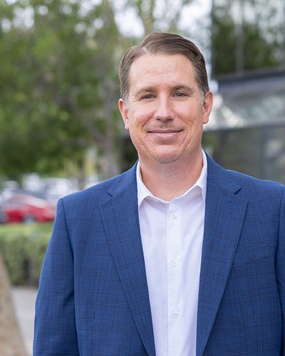 A man wearing a blue suit jacket and white shirt stands outdoors, smiling slightly—exuding executive leadership. Trees and blurred buildings are visible in the background.