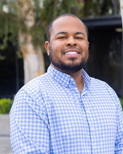 A man with a beard smiles at the camera, embodying executive leadership. He wears a light blue and white checkered button-up shirt and stands outdoors, framed by trees and greenery in the background.