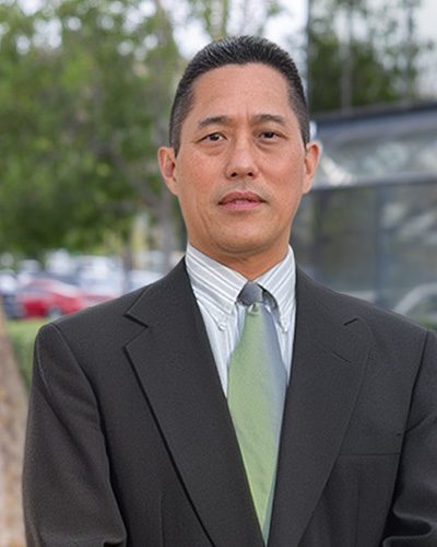 A man exemplifying executive leadership wears a dark suit, light striped shirt, and green tie as he stands outdoors in front of trees and a building, looking directly at the camera.