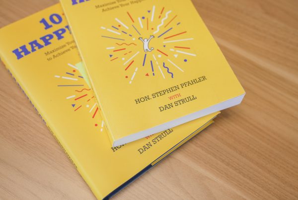 Two bright yellow books titled 10 HAPP, featuring a burst graphic and small white figure on the cover, authored by Hon. Stephen Pfahler with Dan Strull, placed on a wooden surface—perfect for unlocking happiness in your daily life.