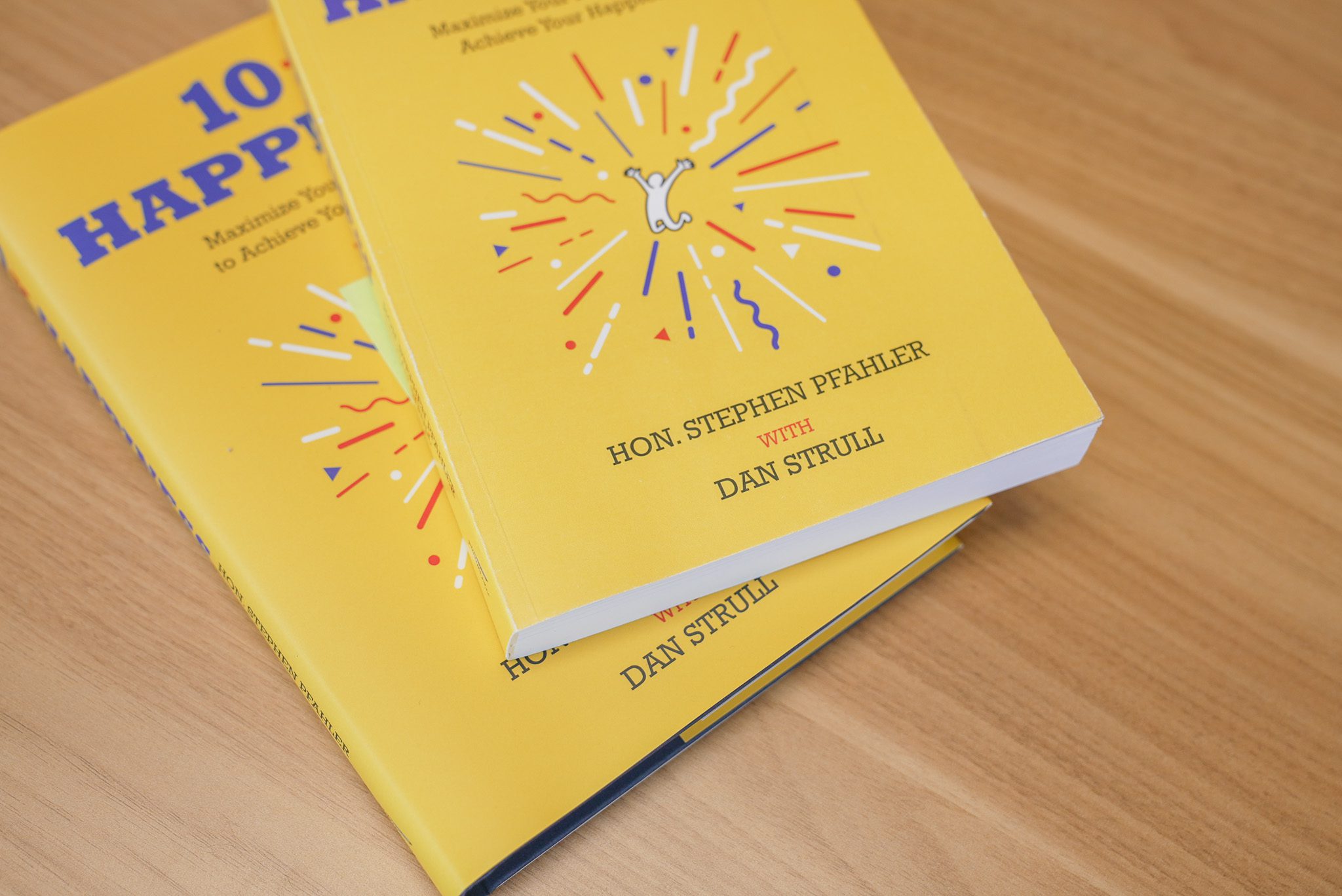 Two bright yellow books titled 10 HAPP, featuring a burst graphic and small white figure on the cover, authored by Hon. Stephen Pfahler with Dan Strull, placed on a wooden surface—perfect for unlocking happiness in your daily life.