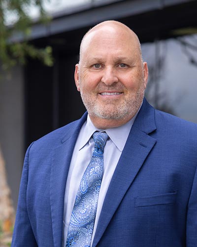 A smiling bald man with a trimmed beard, embodying executive leadership, wears a blue suit, patterned tie, and light blue shirt as he stands outdoors in front of a building.