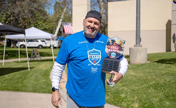 A man in a blue Grateful Hearts T-shirt and black cap stands outdoors holding a Chili Champion trophy, smiling at the camera. Tents, grass, an American flag, and a sign for GoodSuite's ENX Magazine Elite Dealers Award are visible in the background.
