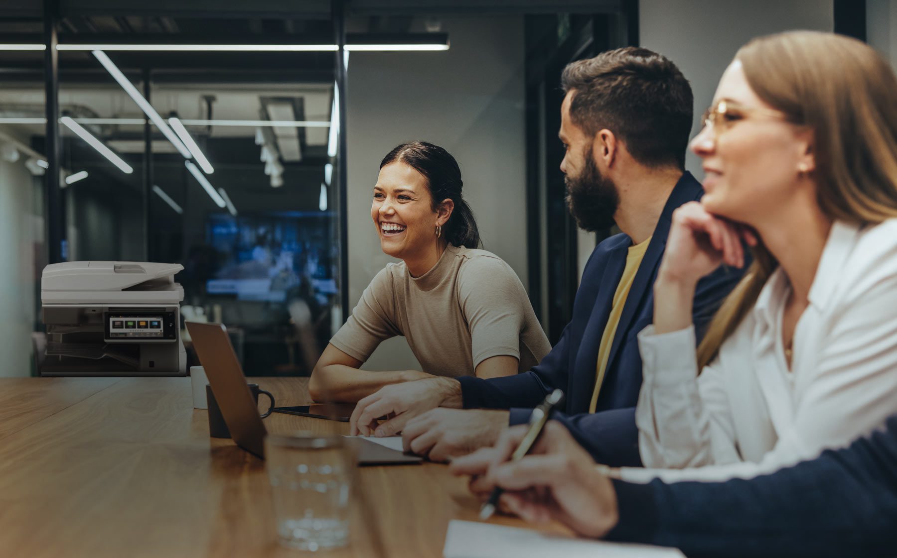 Three colleagues sit at a conference table in an office, engaged in a meeting. One woman smiles brightly, while the other two listen attentively. A printer and glass walls are visible in the background.