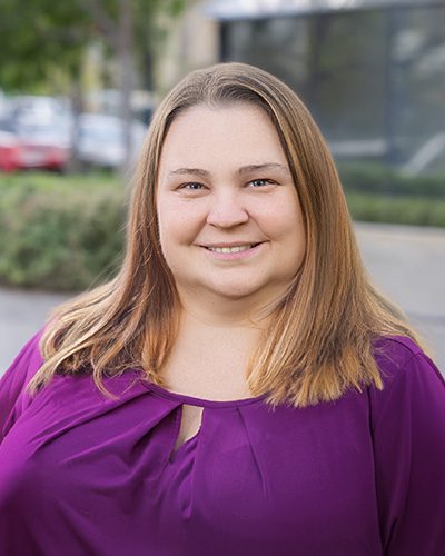 A woman with straight, light brown hair and a purple blouse smiles while standing outdoors; blurred greenery and parked cars are visible in the background, reflecting her approachable executive leadership presence.