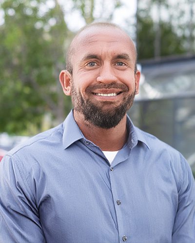 A man with a beard and close-cropped hair smiles at the camera, exuding executive leadership while wearing a light blue button-up shirt, standing outdoors with greenery and blurred buildings in the background.