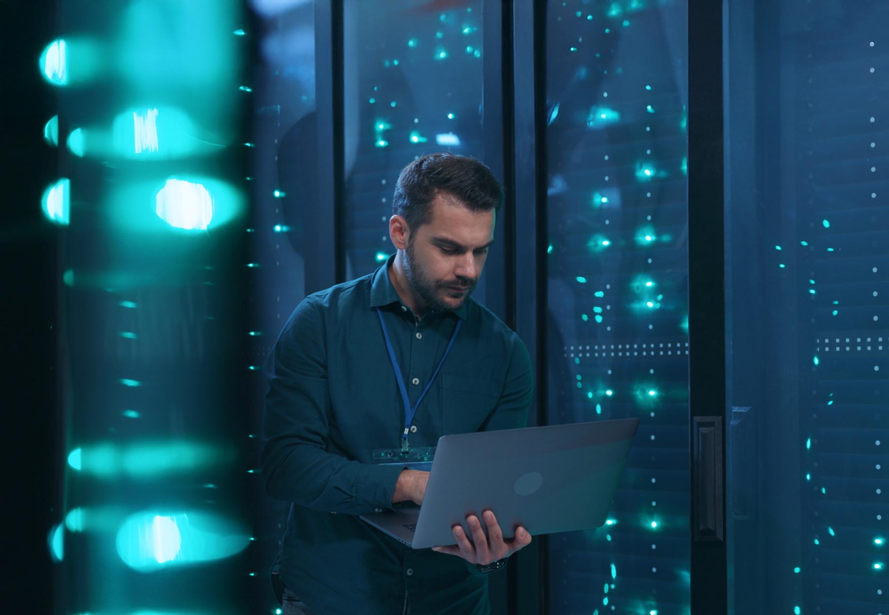 A man stands in a server room, focused on a laptop, surrounded by racks of servers with blue and green indicator lights—a dedicated professional ensuring seamless Managed IT Services. He wears a dark shirt and an ID badge on a lanyard.