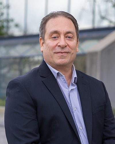 A man with short brown hair wearing a dark suit jacket over a light blue collared shirt stands outdoors, smiling slightly, embodying executive leadership. The background is blurred, showing glass and concrete structures.