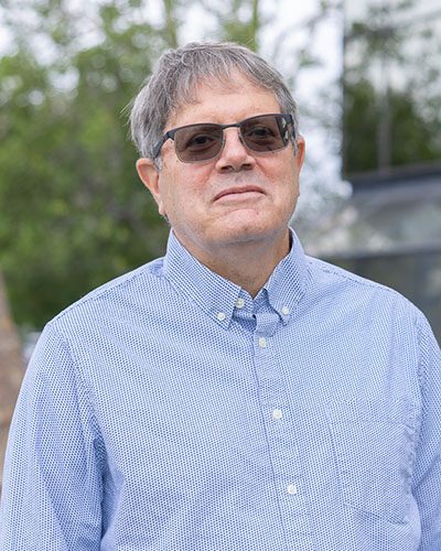 A man with gray hair, embodying executive leadership, wears sunglasses and a light blue checkered button-up shirt as he stands outdoors, with green trees and a building blurred in the background.