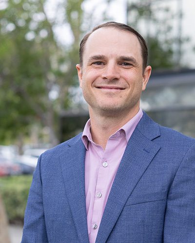 A man wearing a blue suit jacket and light pink shirt smiles at the camera while standing outdoors, exuding executive leadership, with trees and blurred buildings in the background.