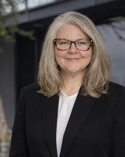 A woman with long gray hair and glasses smiles at the camera, embodying executive leadership. She wears a black blazer over a white blouse, standing outdoors with a blurred background.