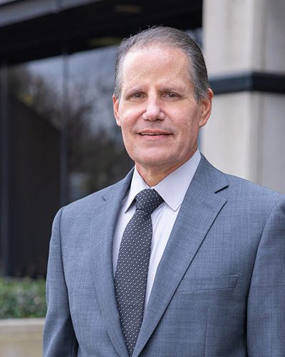 A middle-aged man in a gray suit and polka-dot tie, exemplifying executive leadership, stands outside a modern office building, smiling slightly at the camera.