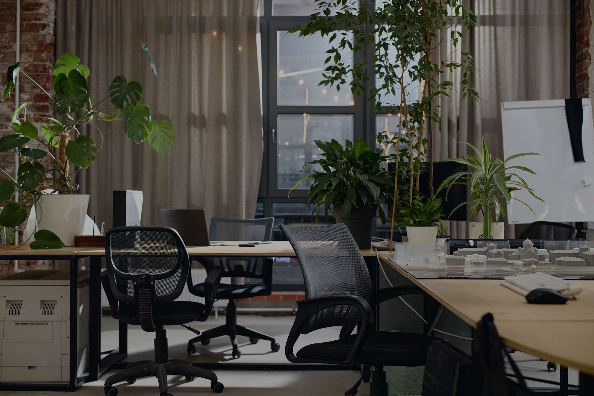 Modern office space with empty desks and chairs, various potted plants, a whiteboard, architectural models, and a GoodSuite copier upgrade. Large window with beige curtains lets in natural light. Ideal for vendor consolidation solutions.