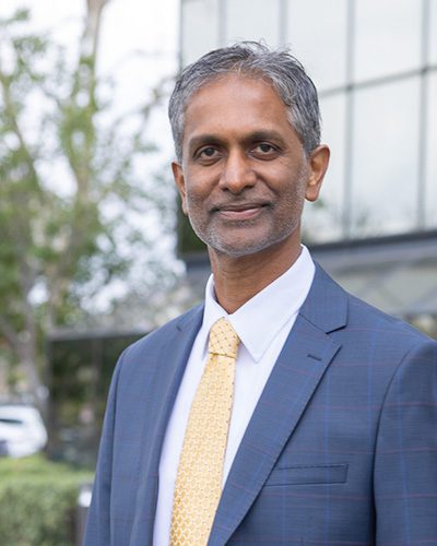 A middle-aged man with short gray hair and a trimmed beard, showcasing executive leadership in a blue suit, white shirt, and yellow tie, stands outdoors in front of a modern glass building.