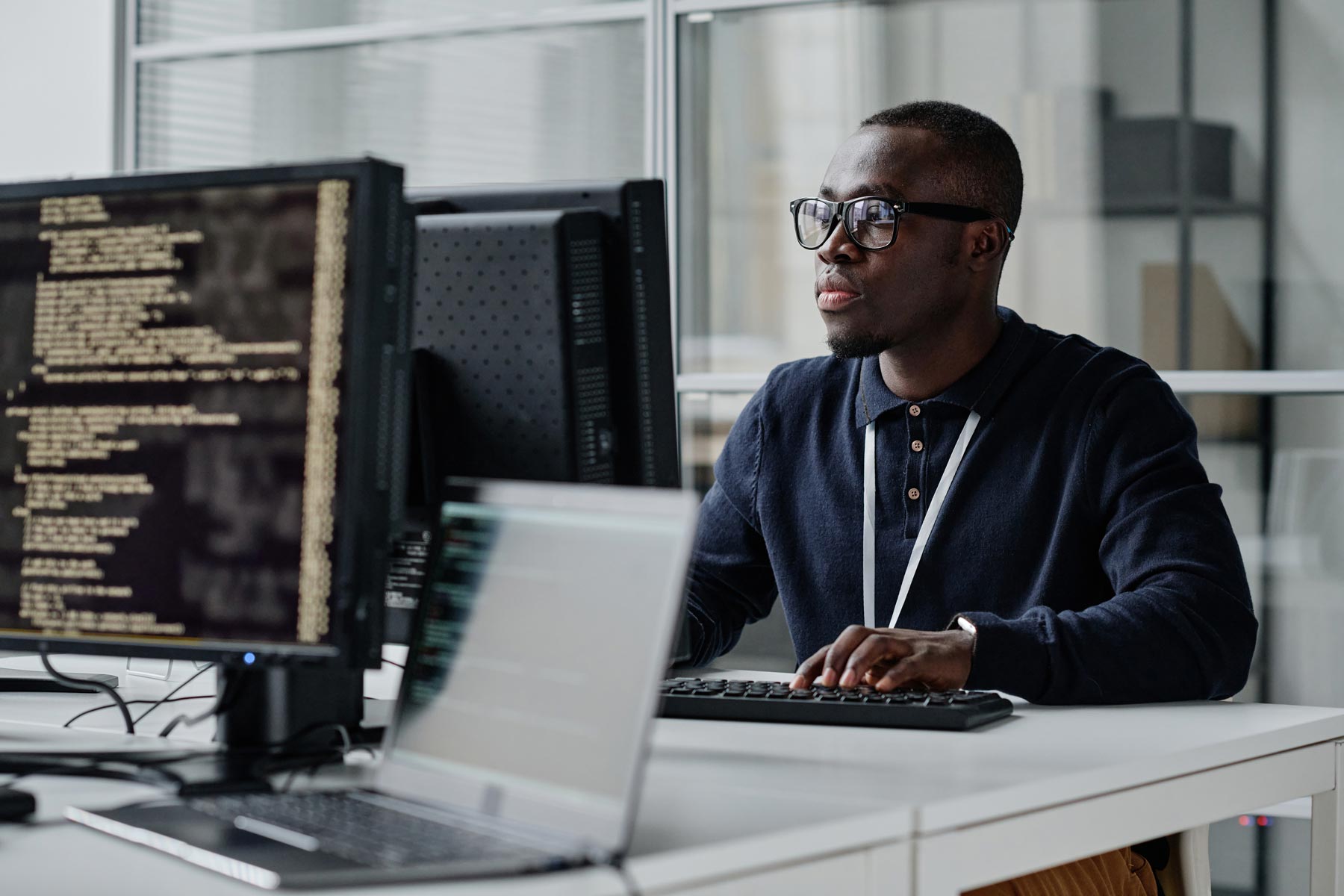 A man wearing glasses and a dark shirt works at a desk with two monitors and a laptop; one monitor displays cybersecurity code. He appears focused in a modern office setting with glass walls.