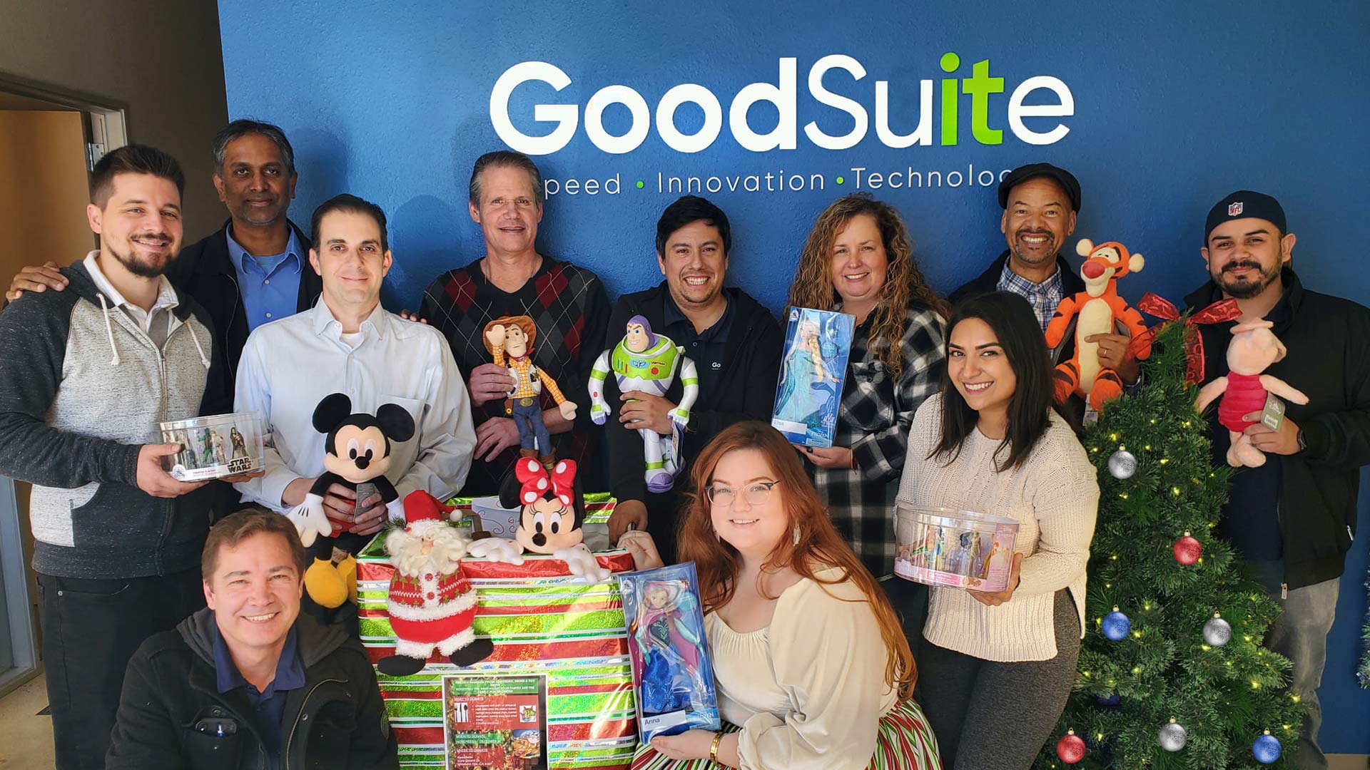 A group of people smiling and holding toys while standing by a decorated Christmas tree and wrapped gifts, with a blue wall behind them displaying the GoodSuite logo.