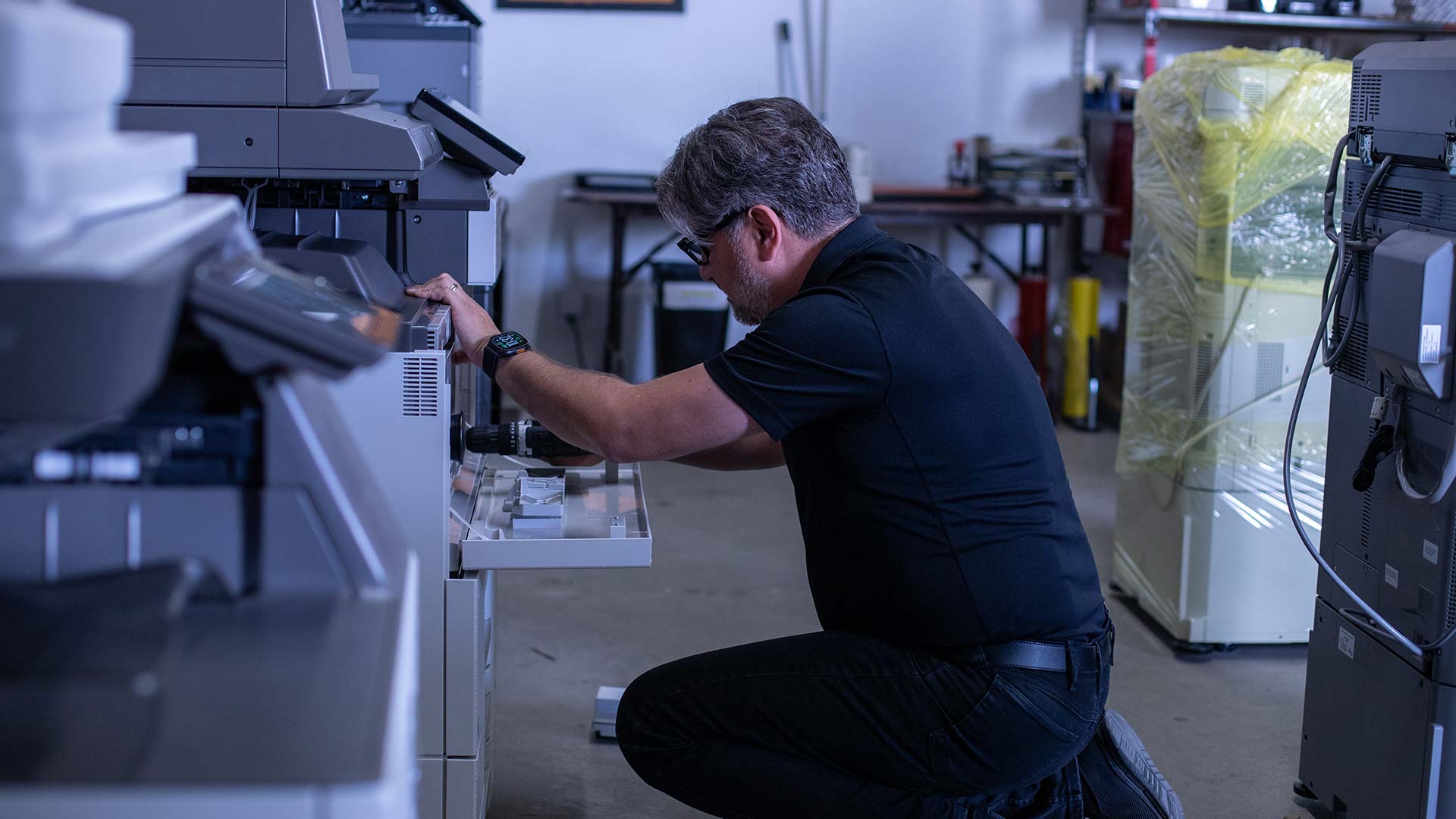 A man wearing glasses and a black shirt kneels on the floor, repairing or inspecting a large printer in an office, showcasing GoodSuite’s dedication to quality service and customer guarantees.