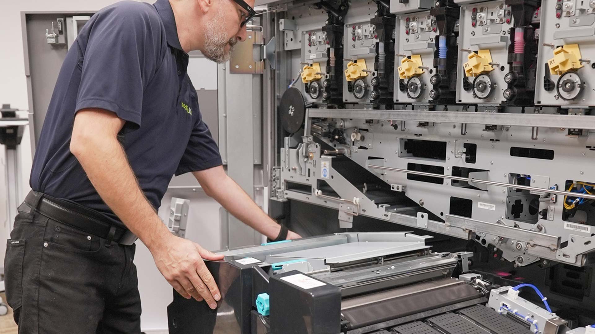 A technician wearing glasses and a navy shirt is servicing a large Production Print industrial printer, with the front panel open, exposing its complex internal components and toner units.