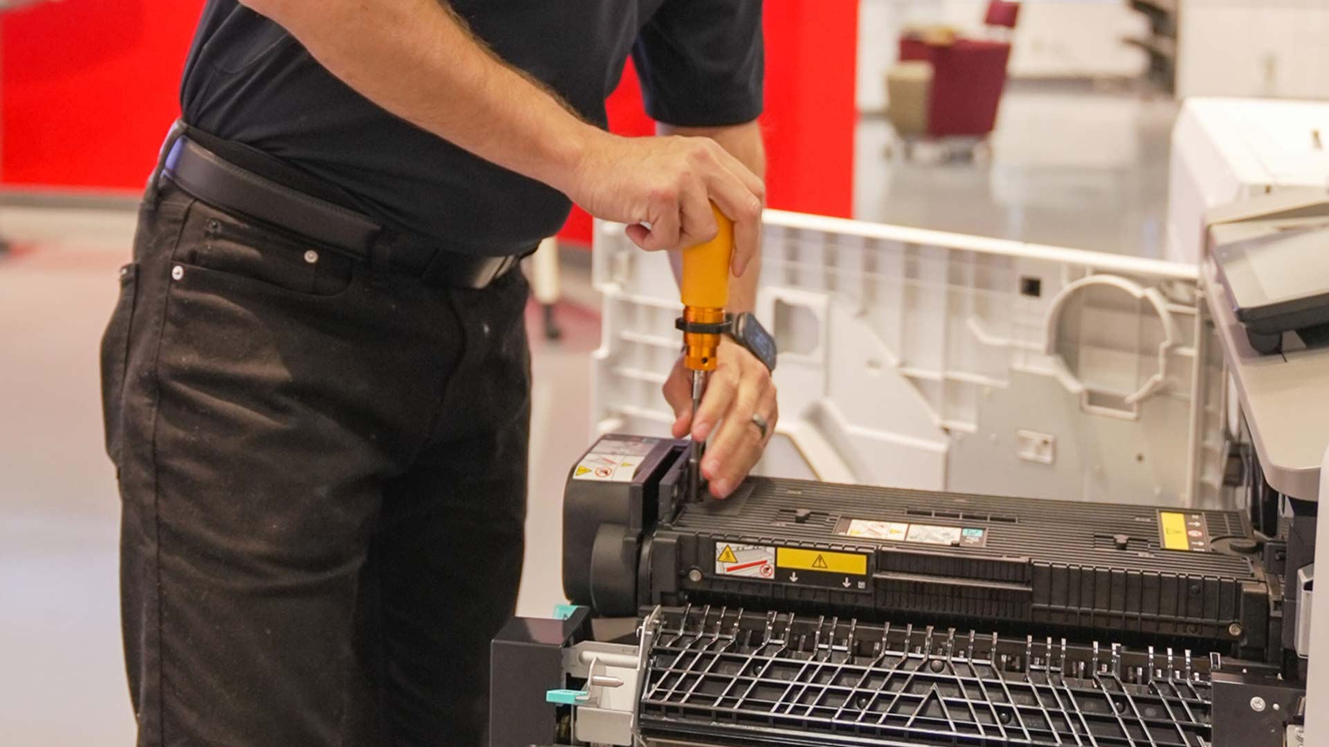 A person using a screwdriver to repair or perform maintenance on an open office printer or copier within designated service areas, with visible internal printer components.