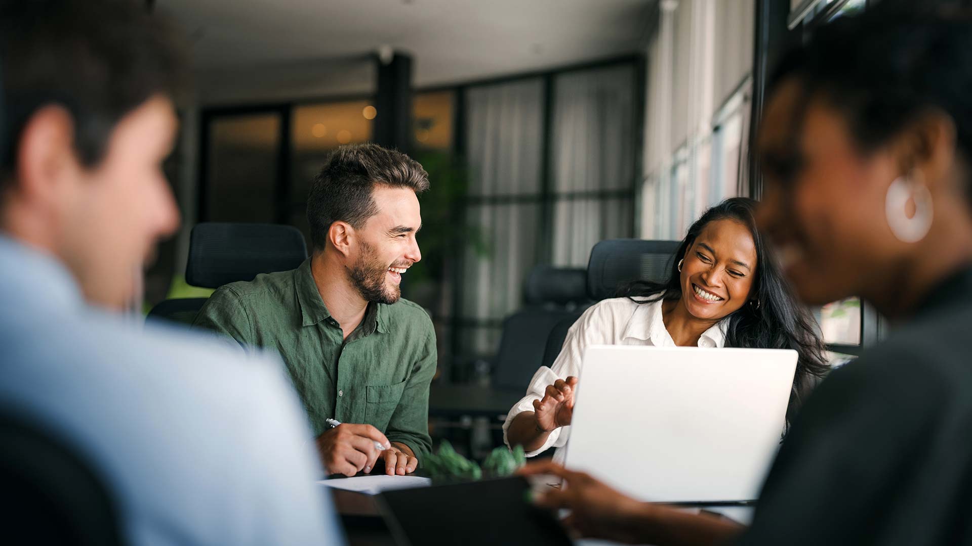Four people sit around a table in a modern office, smiling and talking. Two are focused on a laptop, appearing engaged and happy as they discuss switching to GoodSuite during their collaborative work session.