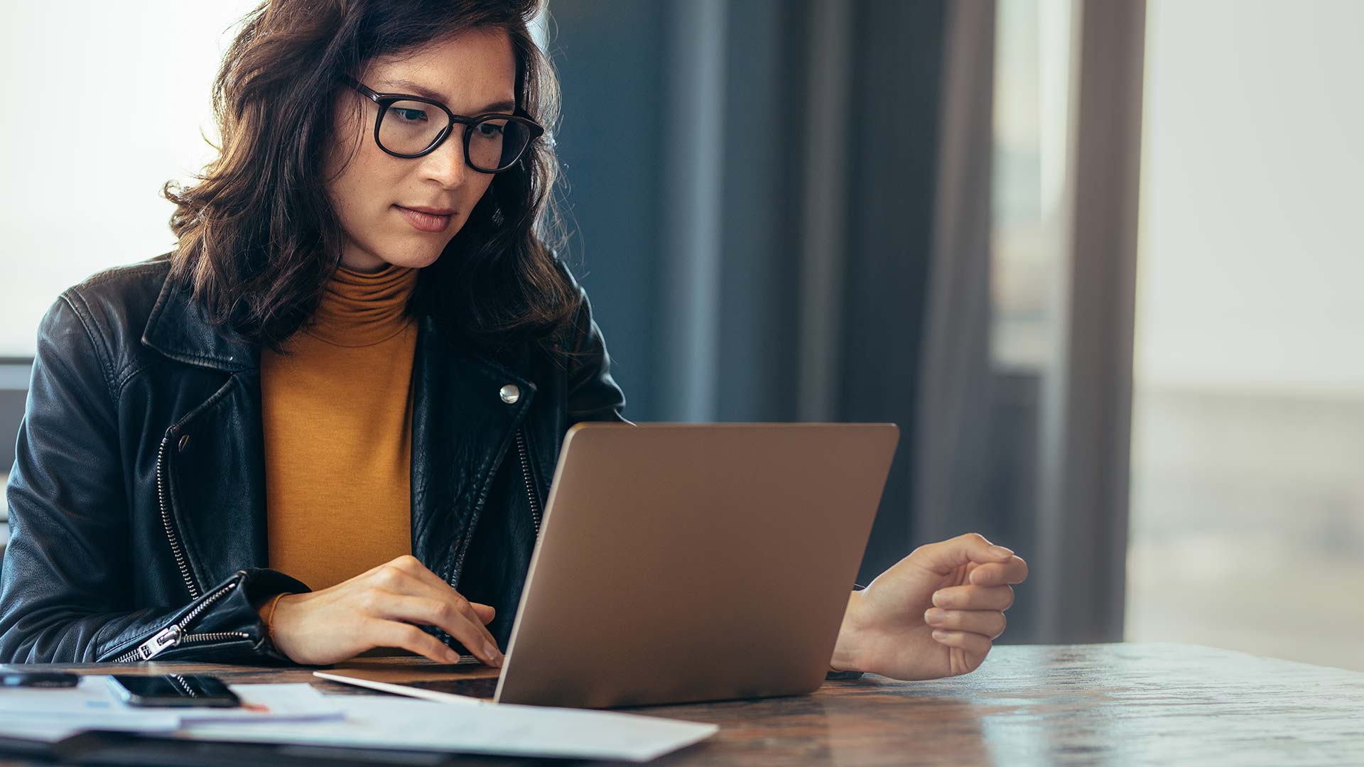 A woman wearing glasses and a black jacket over a mustard turtleneck is working on a laptop at a desk with papers and a pen, in a well-lit room.