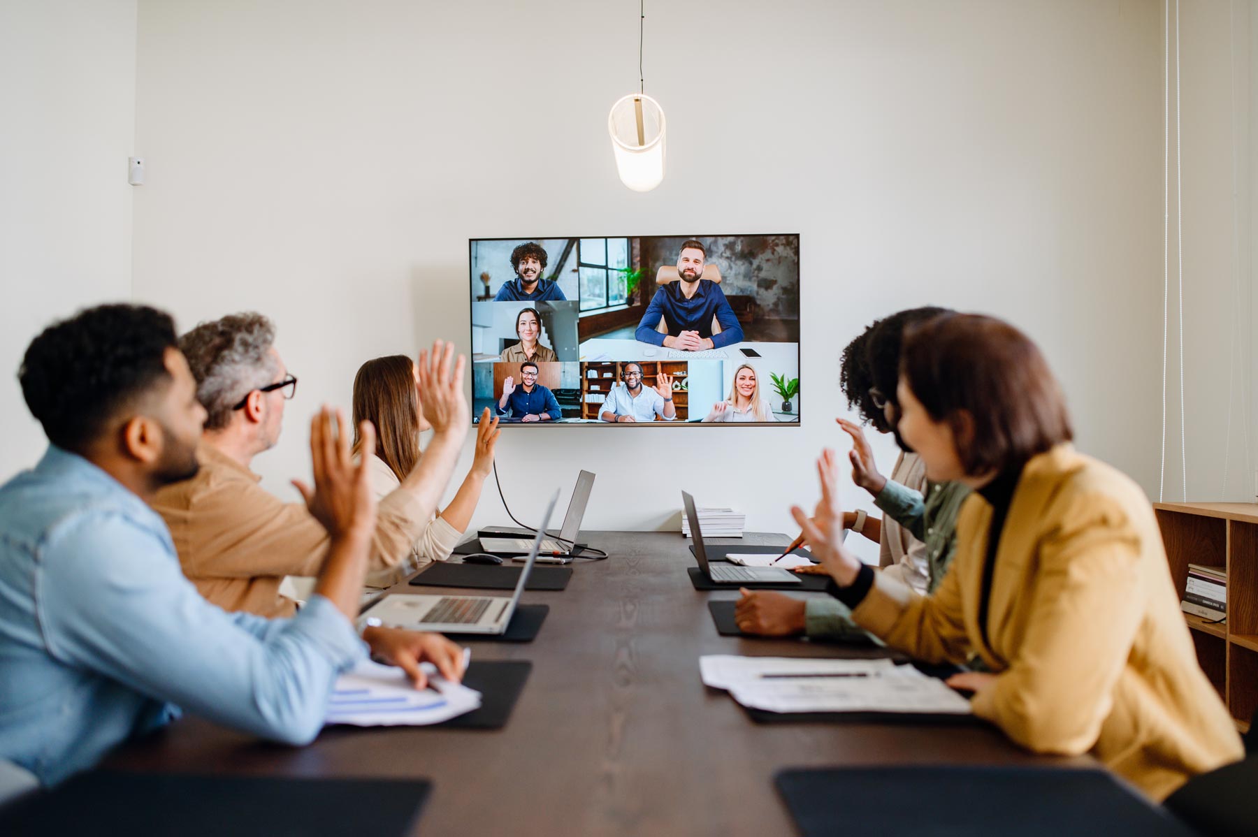 A group of people sitting at a conference table raise their hands while participating in a Unified Communications video call with colleagues displayed on a large screen at the front of the room.