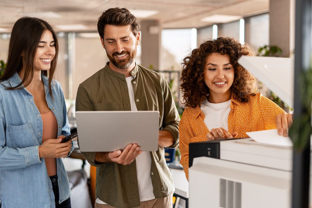 Three young adults smiling in an office collaborate on a project. One woman uses a copier, another holds a phone, and the man in the middle has a laptop—showcasing teamwork enhanced by AI in managed services.