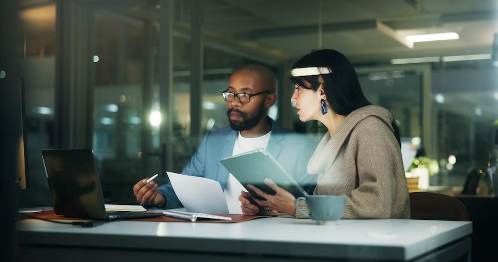 Two people work together at a desk with a laptop and documents, appearing focused on staying CCPA compliant as they discuss strategies in a modern office setting at night.